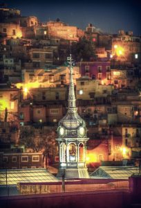 a steeple lit up at night with a hill populated with lots of houses behind it